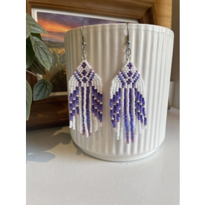 Handmade beaded earrings in shades of purples and white, featuring an intricate geometric pattern and long fringe design. The earrings hang from silver hooks and are displayed against a textured white ceramic background