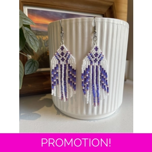 Handmade beaded earrings in shades of purples and white, featuring an intricate geometric pattern and long fringe design. The earrings hang from silver hooks and are displayed against a textured white ceramic background