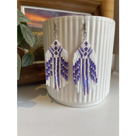 Handmade beaded earrings in shades of purples and white, featuring an intricate geometric pattern and long fringe design. The earrings hang from silver hooks and are displayed against a textured white ceramic background