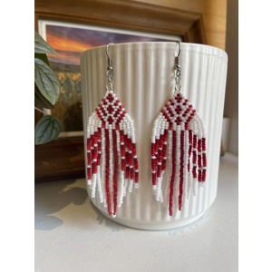 Handmade beaded earrings in shades of reds and white, featuring an intricate geometric pattern and long fringe design. The earrings hang from silver hooks and are displayed against a textured white ceramic background