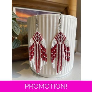Handmade beaded earrings in shades of reds and white, featuring an intricate geometric pattern and long fringe design. The earrings hang from silver hooks and are displayed against a textured white ceramic background