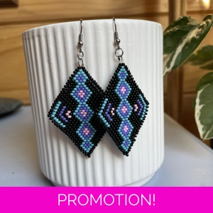 A pair of intricately beaded earrings in a diamond shape, featuring a black background with geometric patterns in shades of blue, purple, and teal. The earrings hang from silver hooks and are displayed against a ribbed white ceramic cup. A green leafy plant is partially visible in the blurred background.