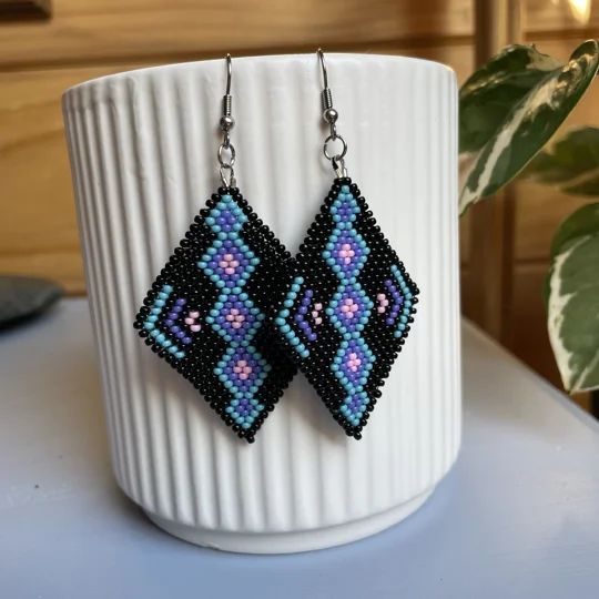 A pair of intricately beaded earrings in a diamond shape, featuring a black background with geometric patterns in shades of blue, purple, and teal. The earrings hang from silver hooks and are displayed against a ribbed white ceramic cup. A green leafy plant is partially visible in the blurred background.