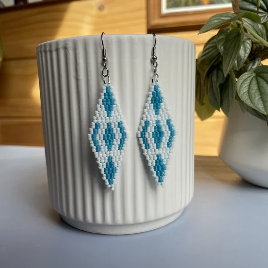 A pair of intricately beaded earrings in a diamond shape, featuring a black background with geometric patterns in shades of blue and white. The earrings hang from silver hooks and are displayed against a ribbed white ceramic cup. A green leafy plant is partially visible in the blurred background.