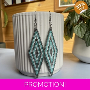 A pair of intricately beaded earrings in a diamond shape, featuring a black background with geometric patterns in shades of grey, turquoise, and white. The earrings hang from silver hooks and are displayed against a ribbed white ceramic cup. A green leafy plant is partially visible in the blurred background.
