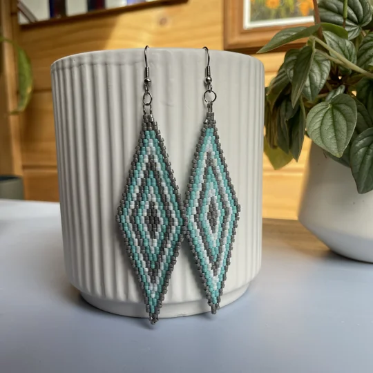 A pair of intricately beaded earrings in a diamond shape, featuring a black background with geometric patterns in shades of grey, turquoise, and white. The earrings hang from silver hooks and are displayed against a ribbed white ceramic cup. A green leafy plant is partially visible in the blurred background.