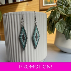 A pair of intricately beaded earrings in a diamond shape, featuring a black background with geometric patterns in shades of grey, turquoise, and white. The earrings hang from silver hooks and are displayed against a ribbed white ceramic cup. A green leafy plant is partially visible in the blurred background.