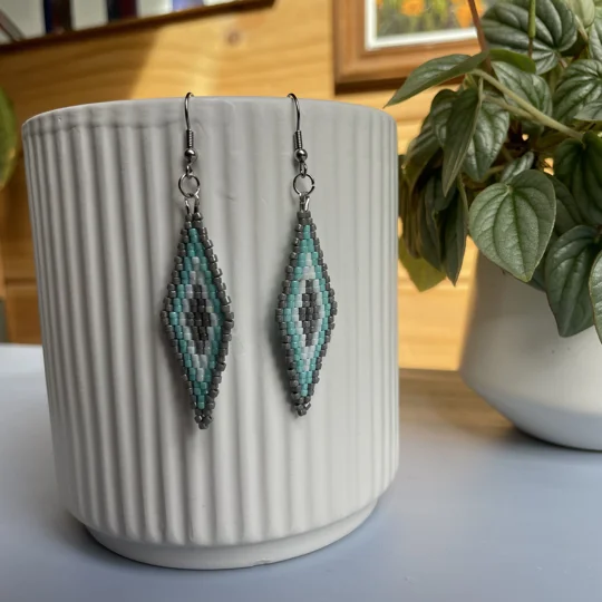 A pair of intricately beaded earrings in a diamond shape, featuring a black background with geometric patterns in shades of grey, turquoise, and white. The earrings hang from silver hooks and are displayed against a ribbed white ceramic cup. A green leafy plant is partially visible in the blurred background.