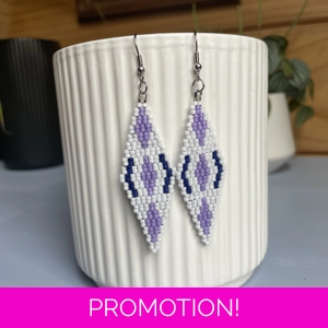 A pair of intricately beaded earrings in a diamond shape, featuring a black background with geometric patterns in shades of purple, and white. The earrings hang from silver hooks and are displayed against a ribbed white ceramic cup. A green leafy plant is partially visible in the blurred background.