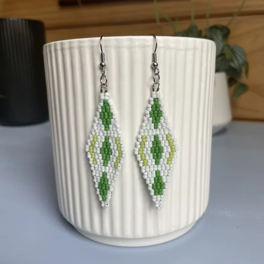 A pair of intricately beaded earrings in a diamond shape, featuring a black background with geometric patterns in shades of green and white. The earrings hang from silver hooks and are displayed against a ribbed white ceramic cup. A green leafy plant is partially visible in the blurred background.