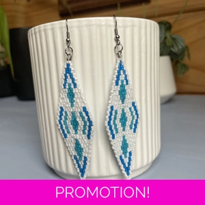 A pair of intricately beaded earrings in a diamond shape, featuring a black background with geometric patterns in shades of blue, and white. The earrings hang from silver hooks and are displayed against a ribbed white ceramic cup. A green leafy plant is partially visible in the blurred background.
