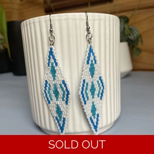 A pair of intricately beaded earrings in a diamond shape, featuring a black background with geometric patterns in shades of blue, and white. The earrings hang from silver hooks and are displayed against a ribbed white ceramic cup. A green leafy plant is partially visible in the blurred background.