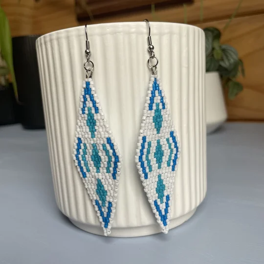 A pair of intricately beaded earrings in a diamond shape, featuring a black background with geometric patterns in shades of blue, and white. The earrings hang from silver hooks and are displayed against a ribbed white ceramic cup. A green leafy plant is partially visible in the blurred background.