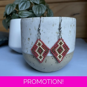 A pair of intricately beaded earrings in a diamond shape, featuring a black background with geometric patterns in shades of Mattee red, beige, and gold colored beads. The earrings hang from silver hooks and are displayed against a ribbed white ceramic cup. A green leafy plant is partially visible in the blurred background.