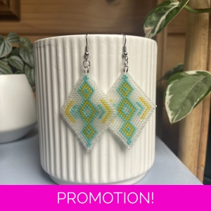 A pair of intricately beaded earrings in a diamond shape, featuring a black background with geometric patterns in shades of frosted white, aqua, and yellow. The earrings hang from silver hooks and are displayed against a ribbed white ceramic cup. A green leafy plant is partially visible in the blurred background.