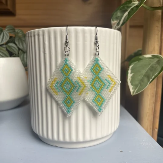 A pair of intricately beaded earrings in a diamond shape, featuring a black background with geometric patterns in shades of frosted white, aqua, and yellow. The earrings hang from silver hooks and are displayed against a ribbed white ceramic cup. A green leafy plant is partially visible in the blurred background.