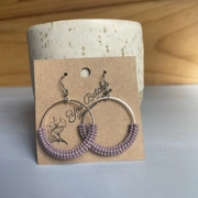 Close-up of handmade bead-wrapped hoop earrings featuring small seed beads intricately woven around a metal hoop, creating a textured, lightweight design in soft neutral tones, displayed on a branded card.