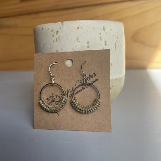 handmade bead-wrapped hoop earrings featuring small seed beads intricately woven around a metal hoop, creating a textured, lightweight design in soft neutral tones, displayed on a branded card. - earthy tones
