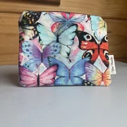 A square, zippered fabric pouch featuring a vibrant pattern of colorful butterflies in shades of blue, purple, pink, and bright red against a soft, mottled background. The pouch has a white side tag that reads 