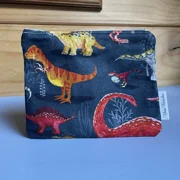 A square, zippered fabric pouch featuring a playful dinosaur pattern on a dark navy blue background. The fabric showcases various illustrated dinosaurs, including a prominent orange and yellow T-Rex, a red Stegosaurus, and a long-necked Brachiosaurus. The pouch features a white side tag reading 