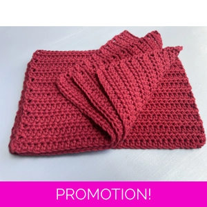A handmade crocheted dishcloth made from red cotton yarn, featuring a textured stitch pattern and soft, neutral tones. The dishcloth is laid flat on a wooden surface.