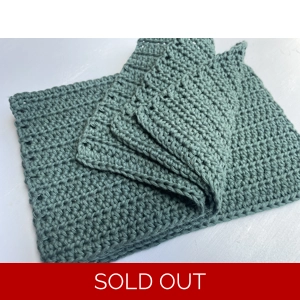 A handmade crocheted dishcloth made from forest green cotton yarn, featuring a textured stitch pattern and soft, neutral tones. The dishcloth is laid flat on a wooden surface.