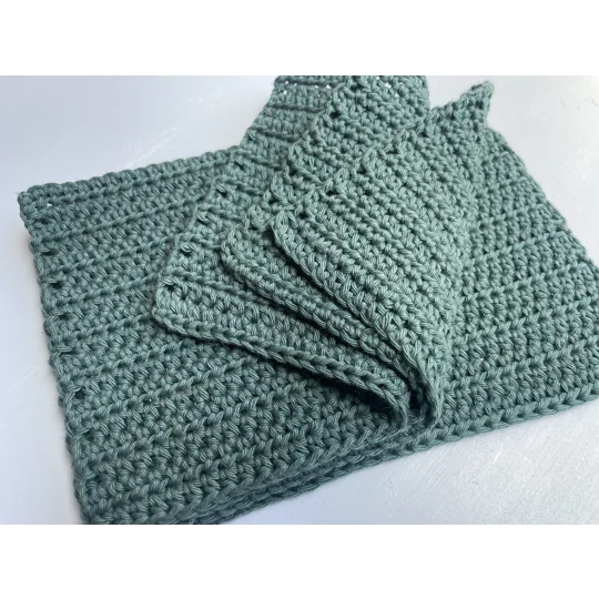 A handmade crocheted dishcloth made from forest green cotton yarn, featuring a textured stitch pattern and soft, neutral tones. The dishcloth is laid flat on a wooden surface.