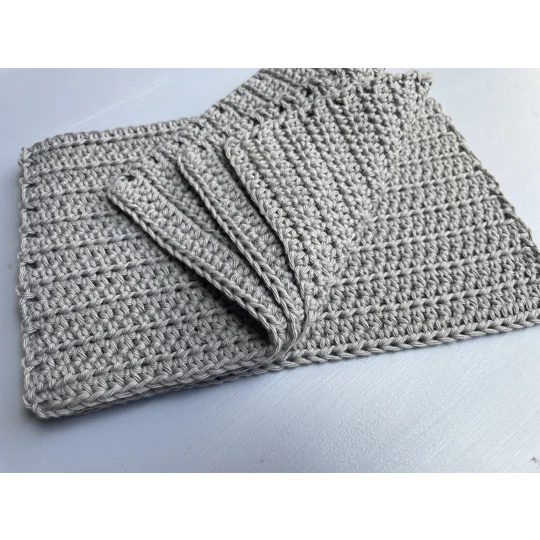A handmade crocheted dishcloth made from grey cotton yarn, featuring a textured stitch pattern and soft, neutral tones. The dishcloth is laid flat on a wooden surface.