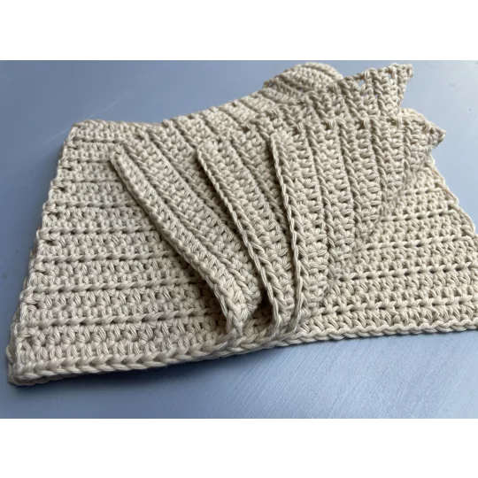 A handmade crocheted dishcloth made from beige cotton yarn, featuring a textured stitch pattern and soft, neutral tones. The dishcloth is laid flat on a wooden surface.