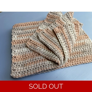 A handmade crocheted dishcloth made from multi hue light orange, and shades of beige cotton yarn, featuring a textured stitch pattern and soft, neutral tones. The dishcloth is laid flat on a wooden surface.