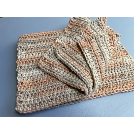 A handmade crocheted dishcloth made from multi hue light orange, and shades of beige cotton yarn, featuring a textured stitch pattern and soft, neutral tones. The dishcloth is laid flat on a wooden surface.