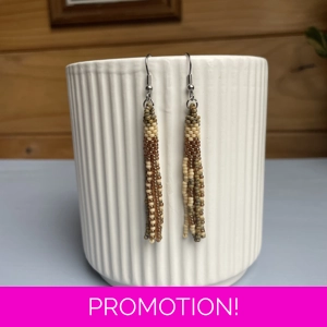 A pair of handmade tube and tassel beaded earrings hanging on a white ribbed ceramic cup. The earrings are made from small glass seed beads in cream, beige, and green, with a stacked tube section at the top and loose beaded tassels hanging below.