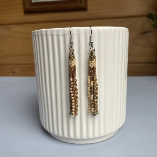 A pair of handmade tube and tassel beaded earrings hanging on a white ribbed ceramic cup. The earrings are made from small glass seed beads in cream, beige, and green, with a stacked tube section at the top and loose beaded tassels hanging below.