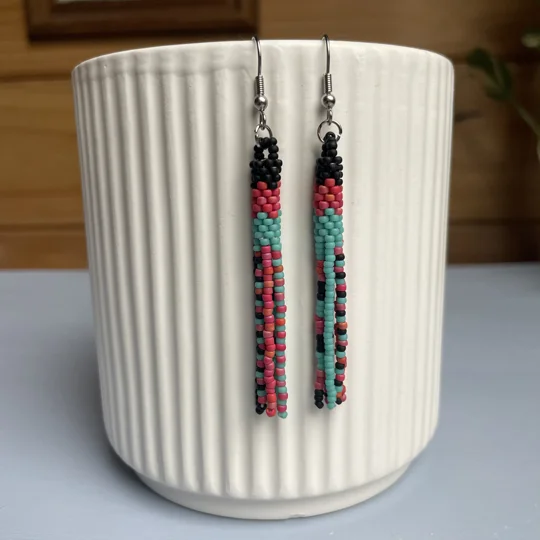 A pair of handmade tube and tassel beaded earrings hanging on a white ribbed ceramic cup. The earrings are made from small glass seed beads in matte coral, matte turquoise, and matte black, with a stacked tube section at the top and loose beaded tassels hanging below.