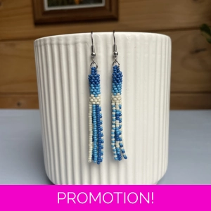 A pair of handmade tube and tassel beaded earrings hanging on a white ribbed ceramic cup. The earrings are made from small glass seed beads in shades of blue, and cream with a stacked tube section at the top and loose beaded tassels hanging below.