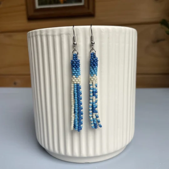 A pair of handmade tube and tassel beaded earrings hanging on a white ribbed ceramic cup. The earrings are made from small glass seed beads in shades of blue, and cream with a stacked tube section at the top and loose beaded tassels hanging below.