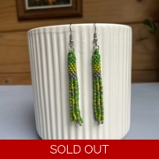 A pair of handmade tube and tassel beaded earrings hanging on a white ribbed ceramic cup. The earrings are made from small glass seed beads in shades of green, and cream with a stacked tube section at the top and loose beaded tassels hanging below.