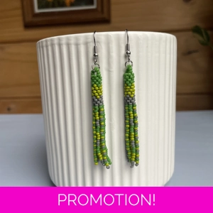 A pair of handmade tube and tassel beaded earrings hanging on a white ribbed ceramic cup. The earrings are made from small glass seed beads in shades of green, and cream with a stacked tube section at the top and loose beaded tassels hanging below.