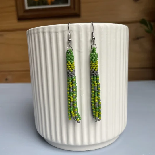 A pair of handmade tube and tassel beaded earrings hanging on a white ribbed ceramic cup. The earrings are made from small glass seed beads in shades of green, and cream with a stacked tube section at the top and loose beaded tassels hanging below.
