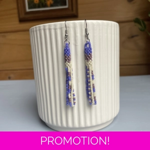 A pair of handmade tube and tassel beaded earrings hanging on a white ribbed ceramic cup. The earrings are made from small glass seed beads in cream, and shades of purple, with a stacked tube section at the top and loose beaded tassels hanging below.