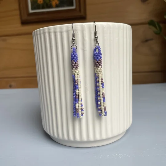 A pair of handmade tube and tassel beaded earrings hanging on a white ribbed ceramic cup. The earrings are made from small glass seed beads in cream, and shades of purple, with a stacked tube section at the top and loose beaded tassels hanging below.