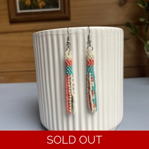 A pair of handmade tube and tassel beaded earrings hanging on a white ribbed ceramic cup. The earrings are made from small glass seed beads in coral, white, and turquoise, with a stacked tube section at the top and loose beaded tassels hanging below.