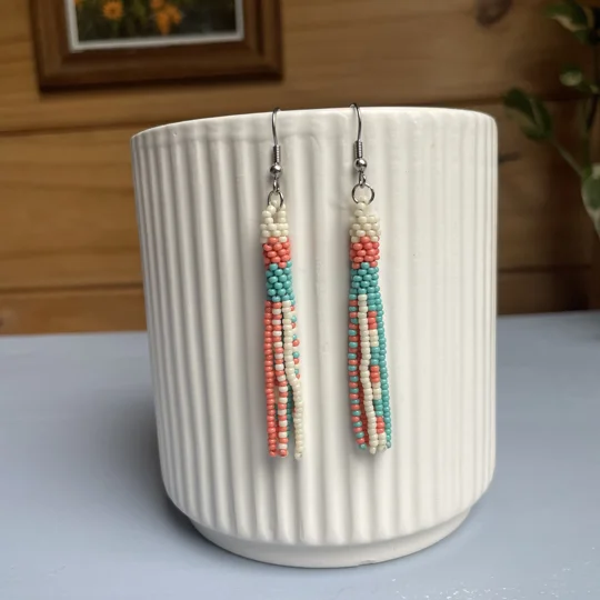 A pair of handmade tube and tassel beaded earrings hanging on a white ribbed ceramic cup. The earrings are made from small glass seed beads in coral, white, and turquoise, with a stacked tube section at the top and loose beaded tassels hanging below.