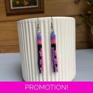 A pair of handmade tube and tassel beaded earrings hanging on a white ribbed ceramic cup. The earrings are made from small glass seed beads in purple, pink, and black, with a stacked tube section at the top and loose beaded tassels hanging below.