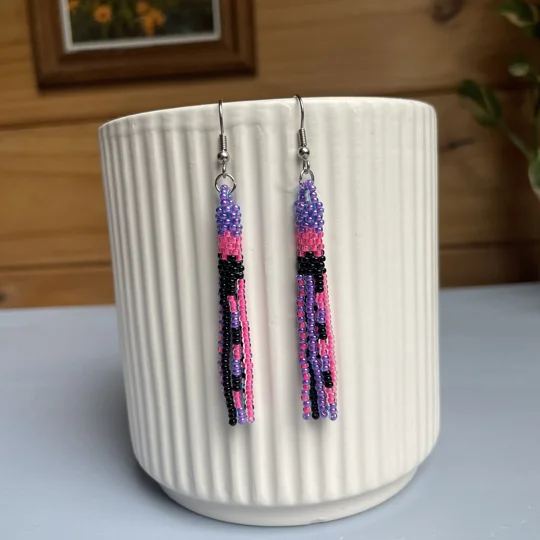 A pair of handmade tube and tassel beaded earrings hanging on a white ribbed ceramic cup. The earrings are made from small glass seed beads in purple, pink, and black, with a stacked tube section at the top and loose beaded tassels hanging below.